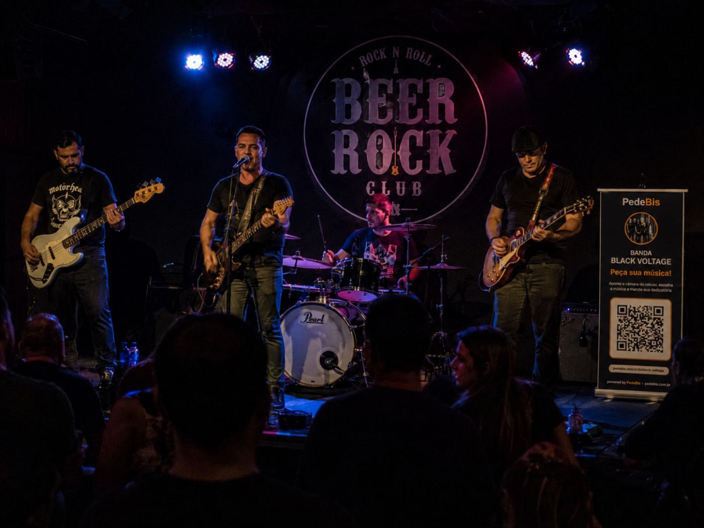 Banda tocando em clube de rock com banner PedeBis no palco
